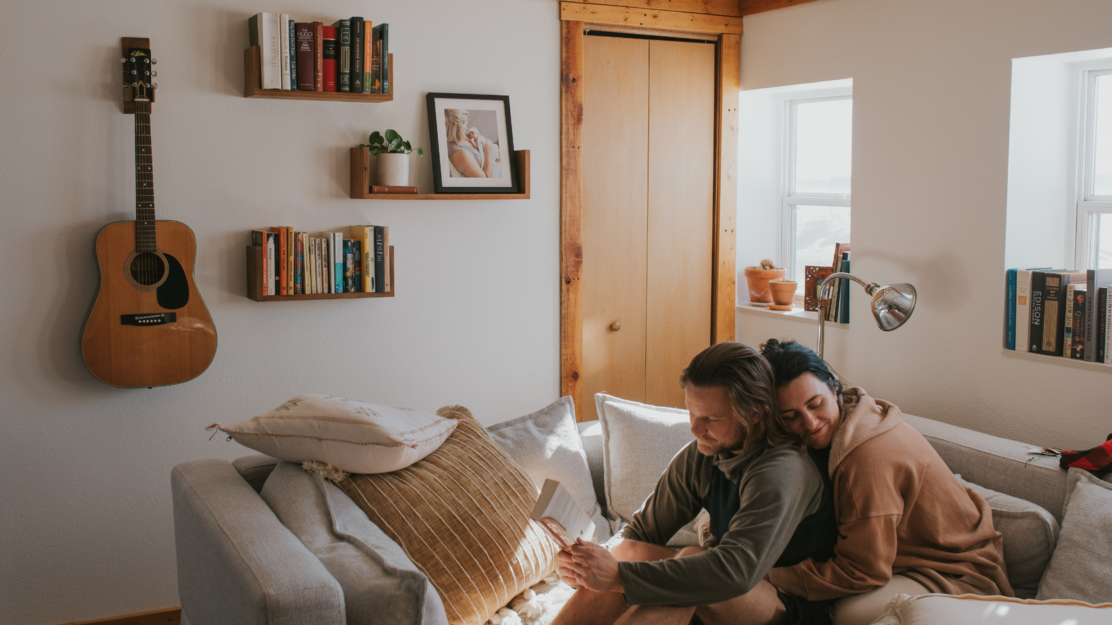 A couple sitting on a couch, one reading a book the other hugging him from behind, with floating u shelves in walnut and a guitar mounted on the wall to the left of them