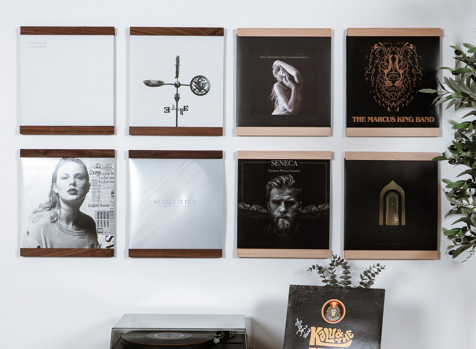 A styled interior wall showing a wall-mounted floating wooden shelf in Walnut and Maple hardwoods that trim the top and bottom of a vinyl record. Eight units organized in a grid above a record player. The product is the wall shelf itself, shown in a real room with wood flooring, a plant, a basket with a throw blanket. 