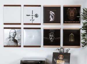 A styled interior wall showing a wall-mounted floating wooden shelf in Walnut and Maple hardwoods that trim the top and bottom of a vinyl record. Eight units organized in a grid above a record player. The product is the wall shelf itself, shown in a real room with wood flooring, a plant, a basket with a throw blanket. 