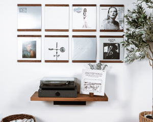 A styled interior wall showing a wall-mounted floating wooden shelf That trims the top and bottom of a vinyl record. Eight units organized in a grid above a record player. The product is the wall shelf itself, shown in a real room with wood flooring, a plant, a basket with a throw blanket. 